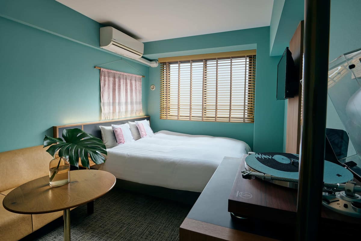 A hotel room with a bed, a small table, a plant, a window with blinds, and a television.