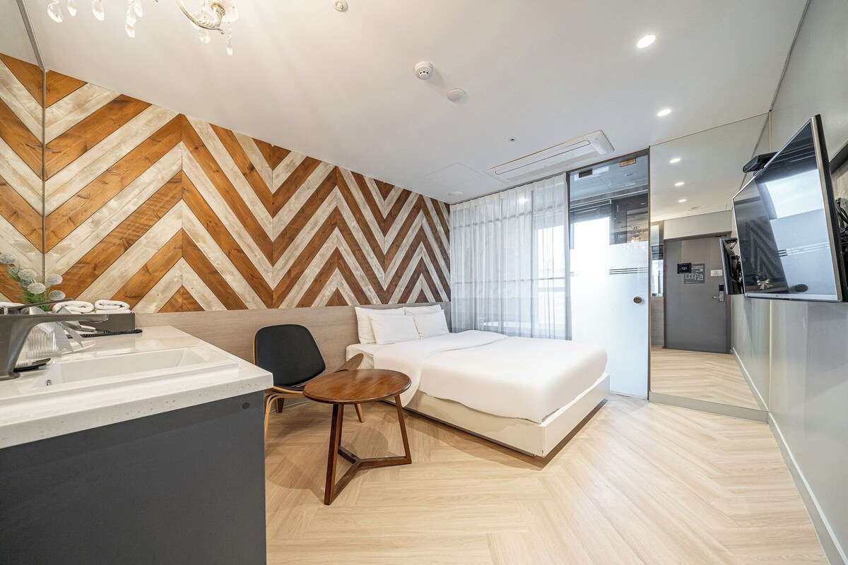 Ein modernes Hotelzimmer mit Wand im Fischgrätmuster, einem Bett, einem kleinen runden Tisch, einem Sessel, einem Waschbecken und Blick auf die Küche.