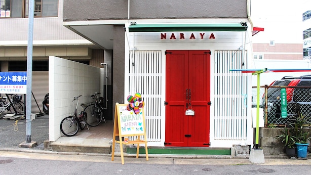 A storefront with a red door and the name 'NARAYA' above it. There is a small easel with a colorful painting in front of the door. - Guest House Naraya - Hostel (Fukuoka)