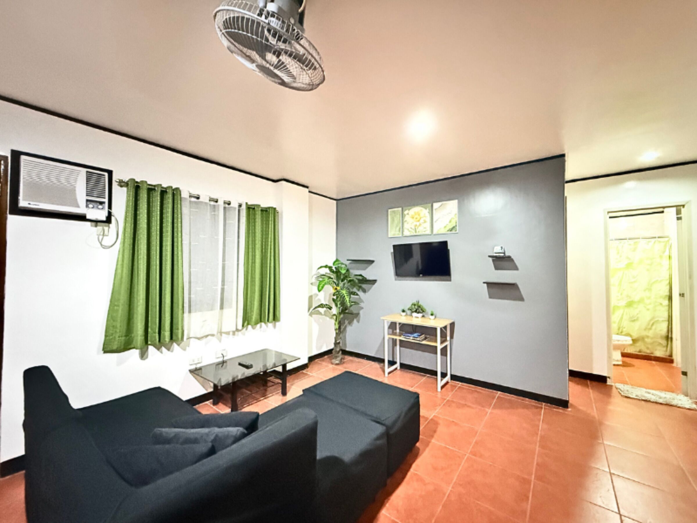 Un salon avec un canapé noir, une petite table, une plante en pot, un téléviseur fixé au mur et un ventilateur.