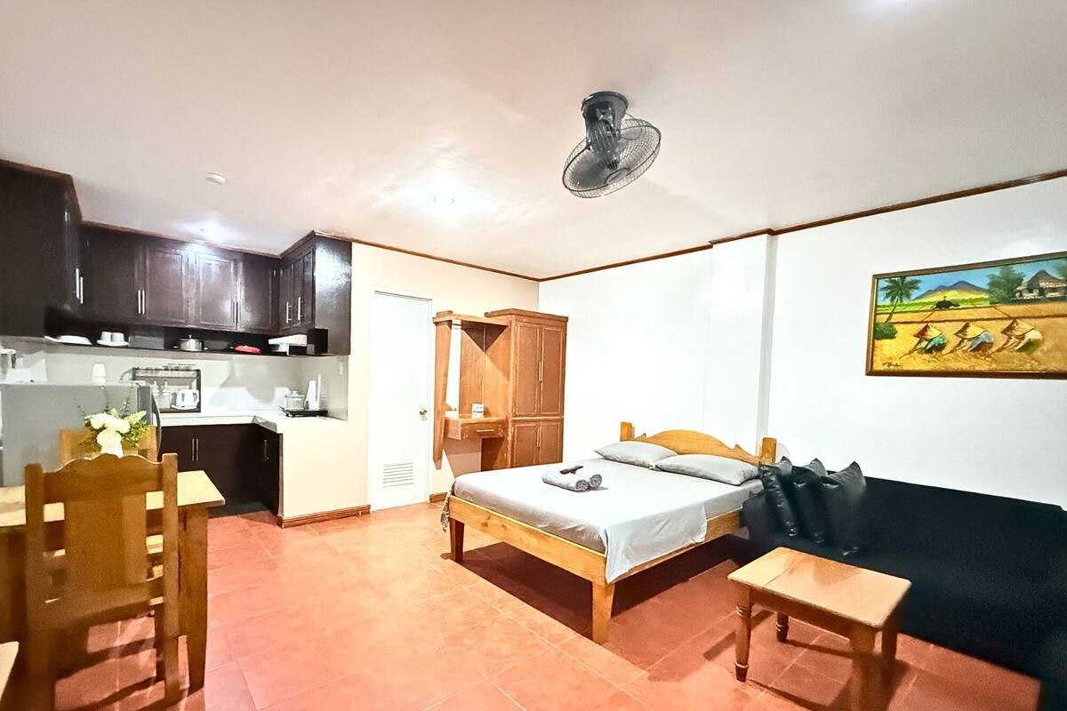 Une pièce de taille modeste comprenant un lit, une petite table, une chaise, un coin cuisine et un ventilateur de plafond.