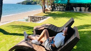 Une personne se détend sur un fauteuil en osier, près d’une plage, avec vue sur l’océan et un mur de pierres.