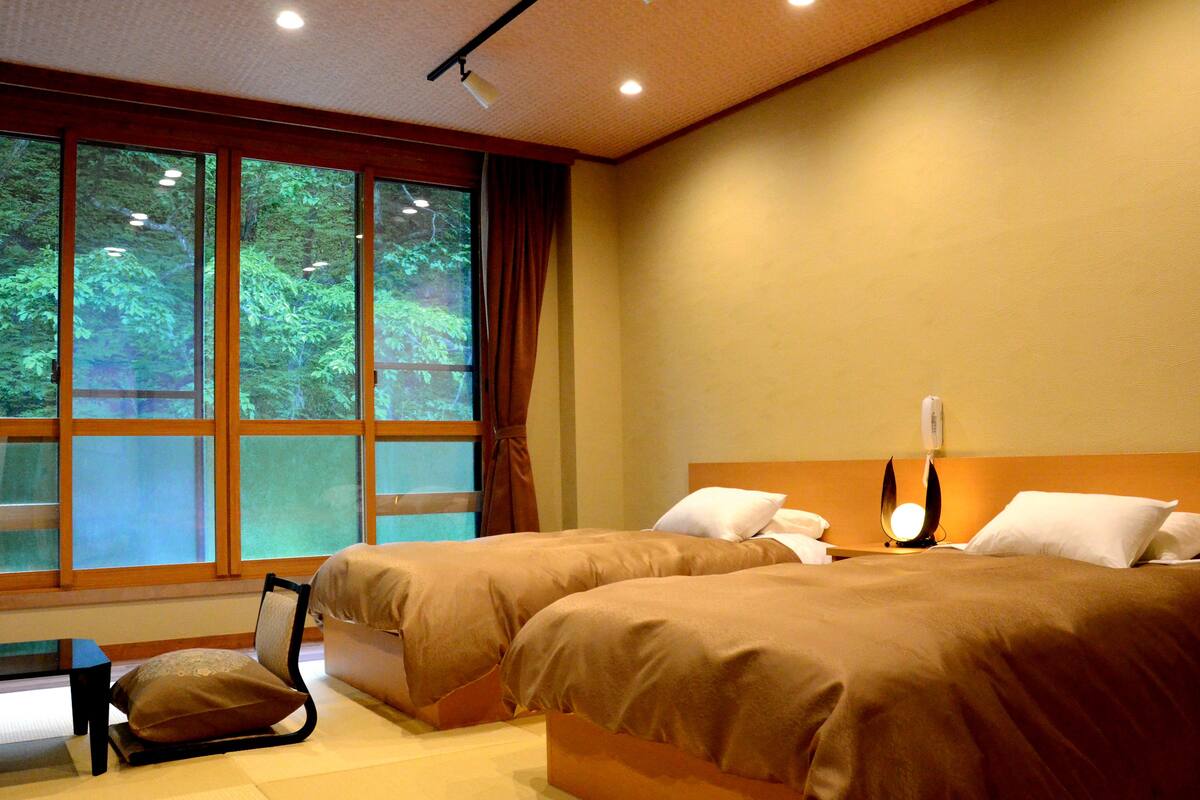 A hotel room with two beds, a bedside table, a chair, and a large window with a view of trees.