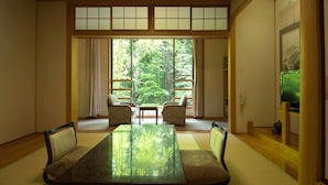 Una habitación japonesa tradicional con suelo de tatami, una mesa baja, dos sillas y un ventanal con vistas a la vegetación.