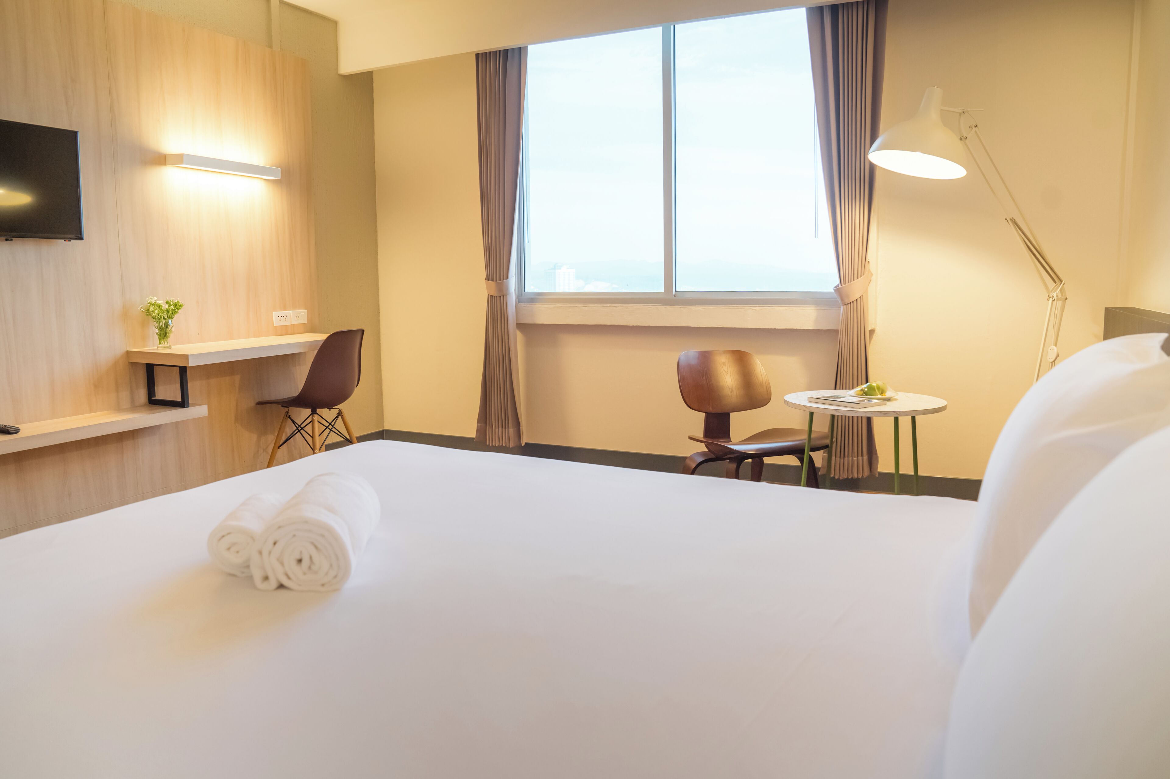 A hotel room with a large bed, a TV mounted on the wall, a small table with a chair, and a window with curtains.