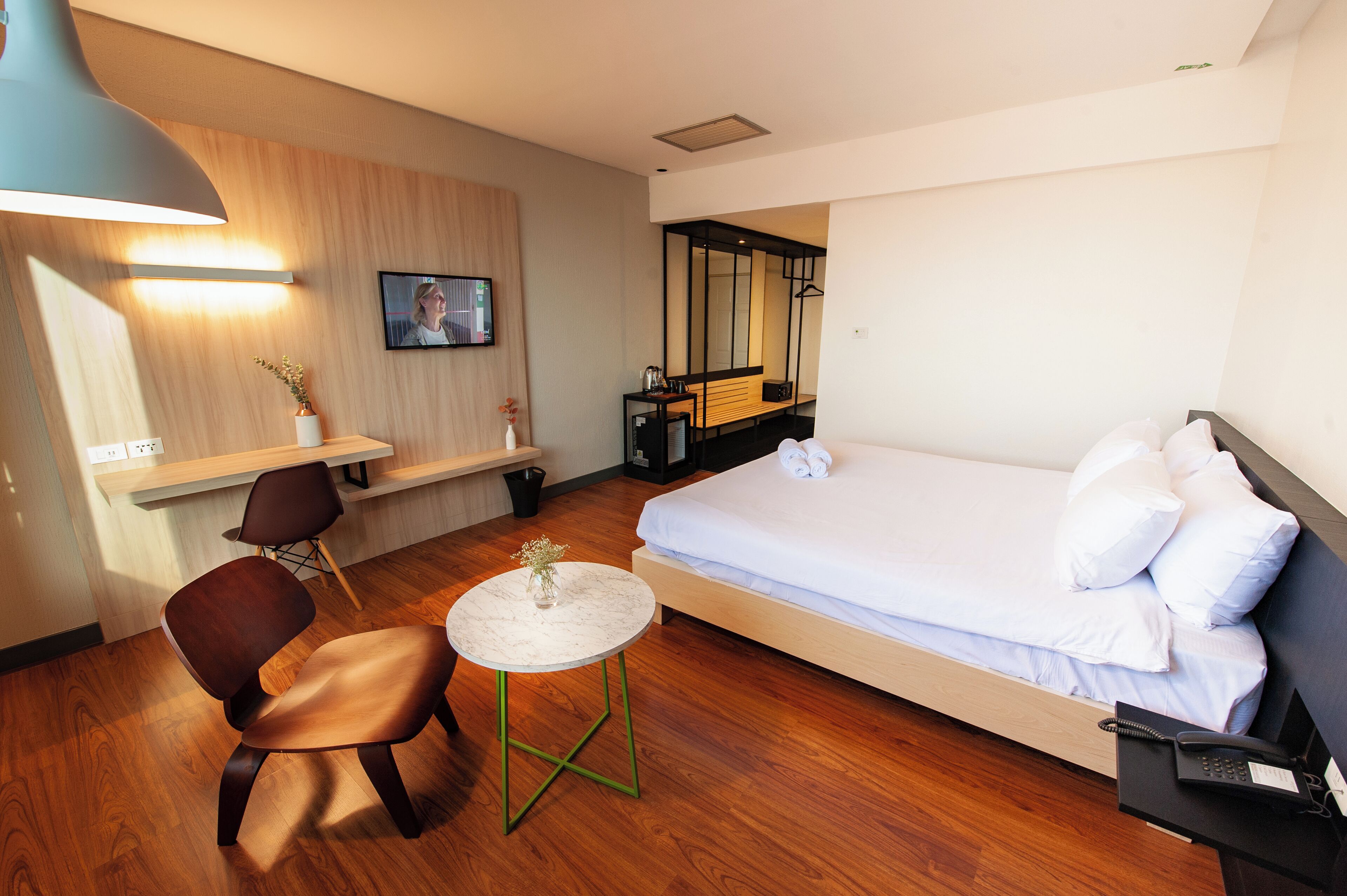 A hotel room with a bed, a desk with a chair, a small table with a vase, and a TV on the wall.