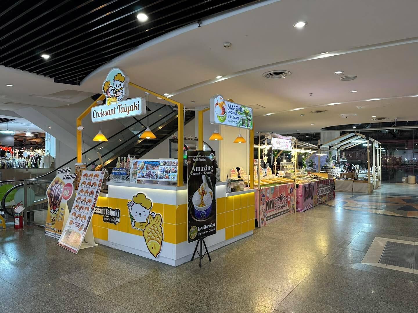 A food stall with a yellow and white color scheme, featuring signs for 'Croissant Taiyaki' and 'Amazing Coconut Ice Cream'.