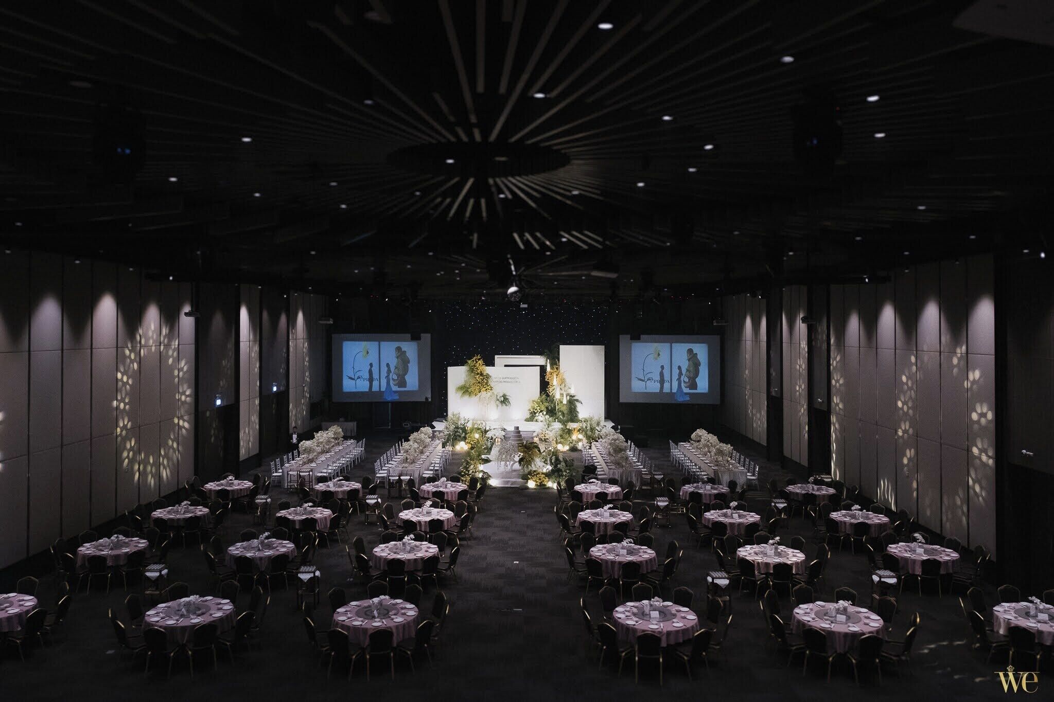 A banquet hall with rows of round tables, a central floral arrangement, and a projection screen at the front.