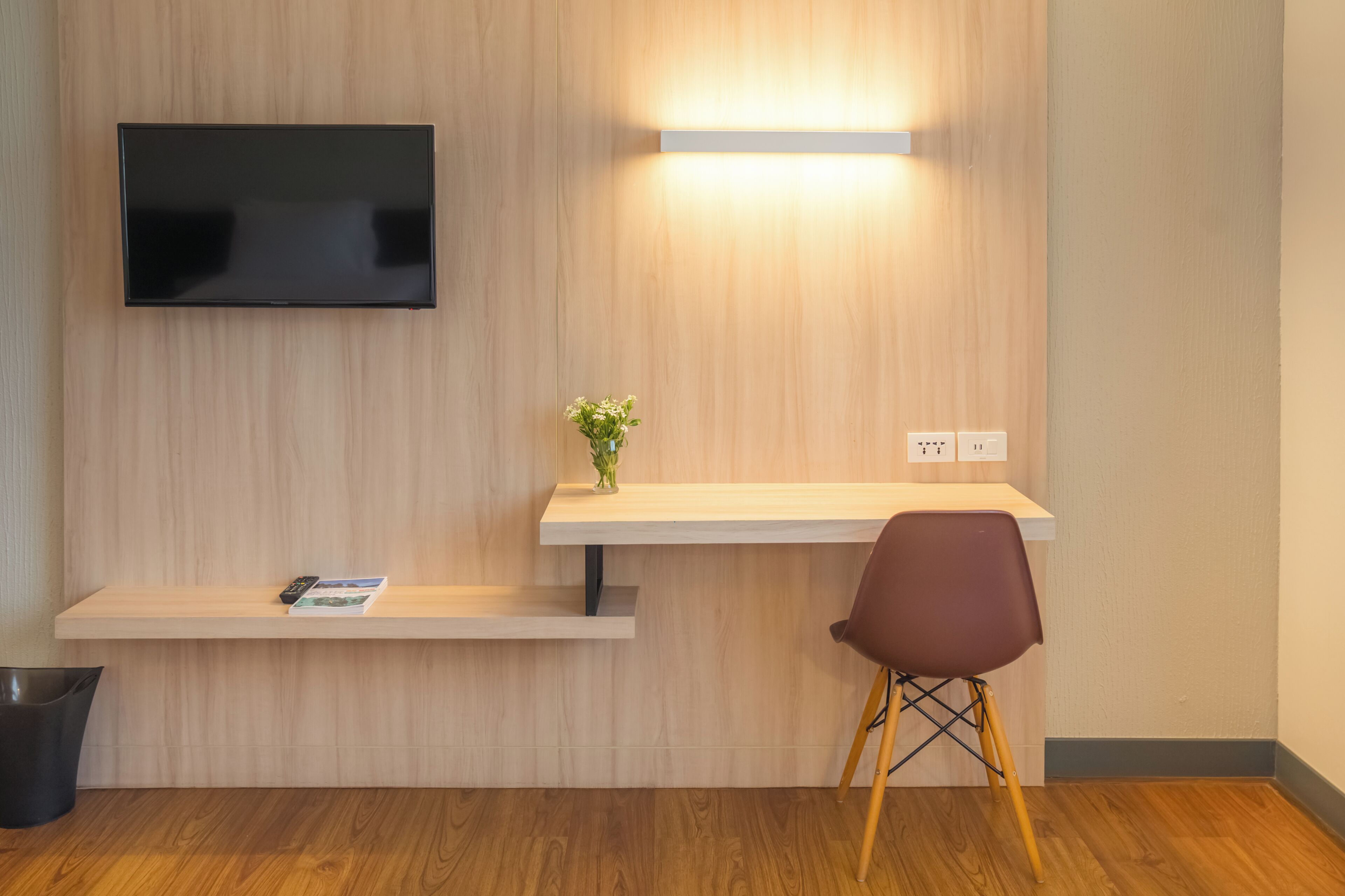 A modern room with a flat-screen TV mounted on the wall, a small desk with a vase of flowers, and a single chair.