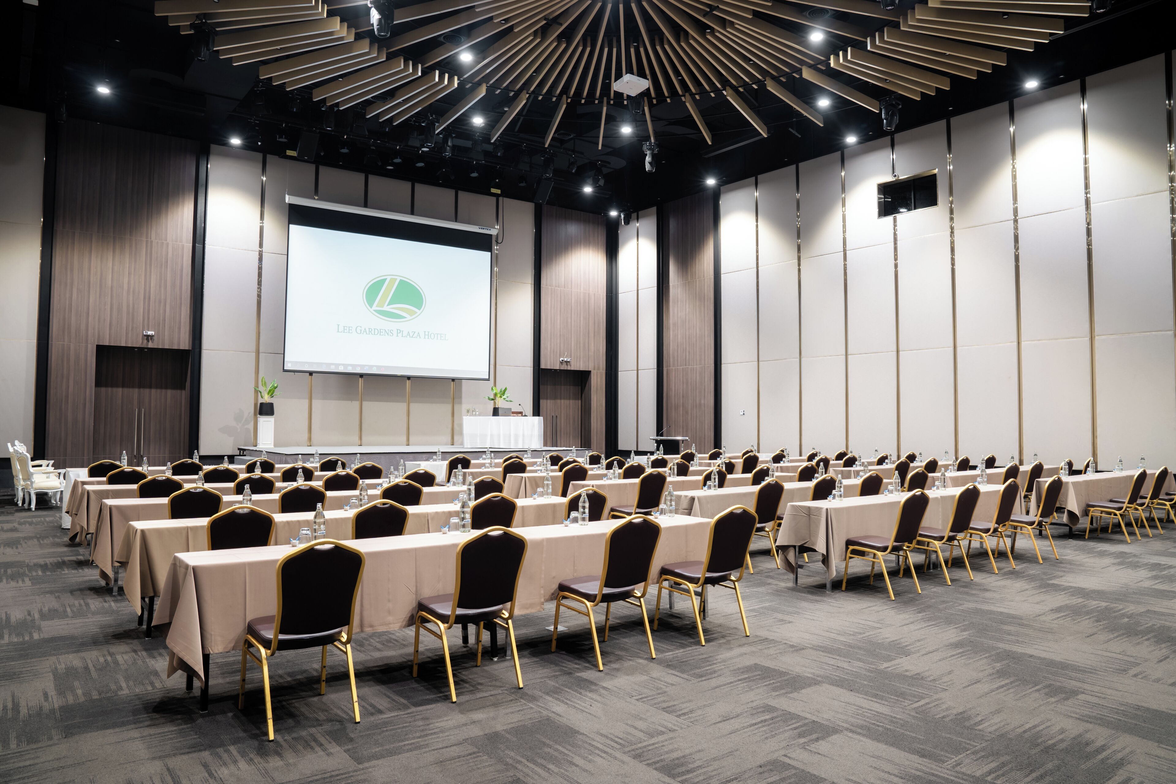Un salón de conferencias con mesas largas, sillas y una pantalla grande que muestra el logotipo del Hotel Lee Gardens Plaza.