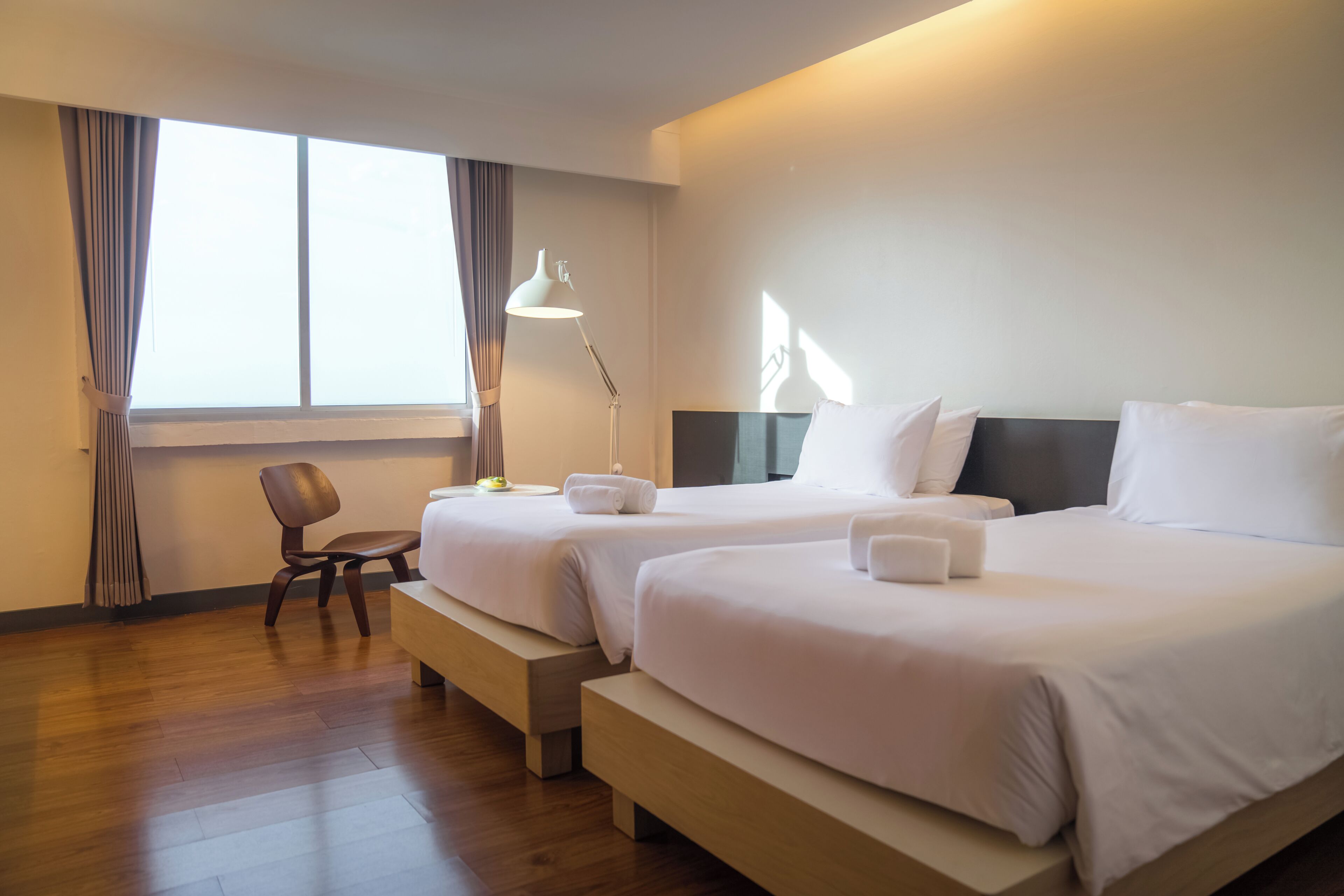 A hotel room with two beds, a chair, a lamp, and a window with curtains.