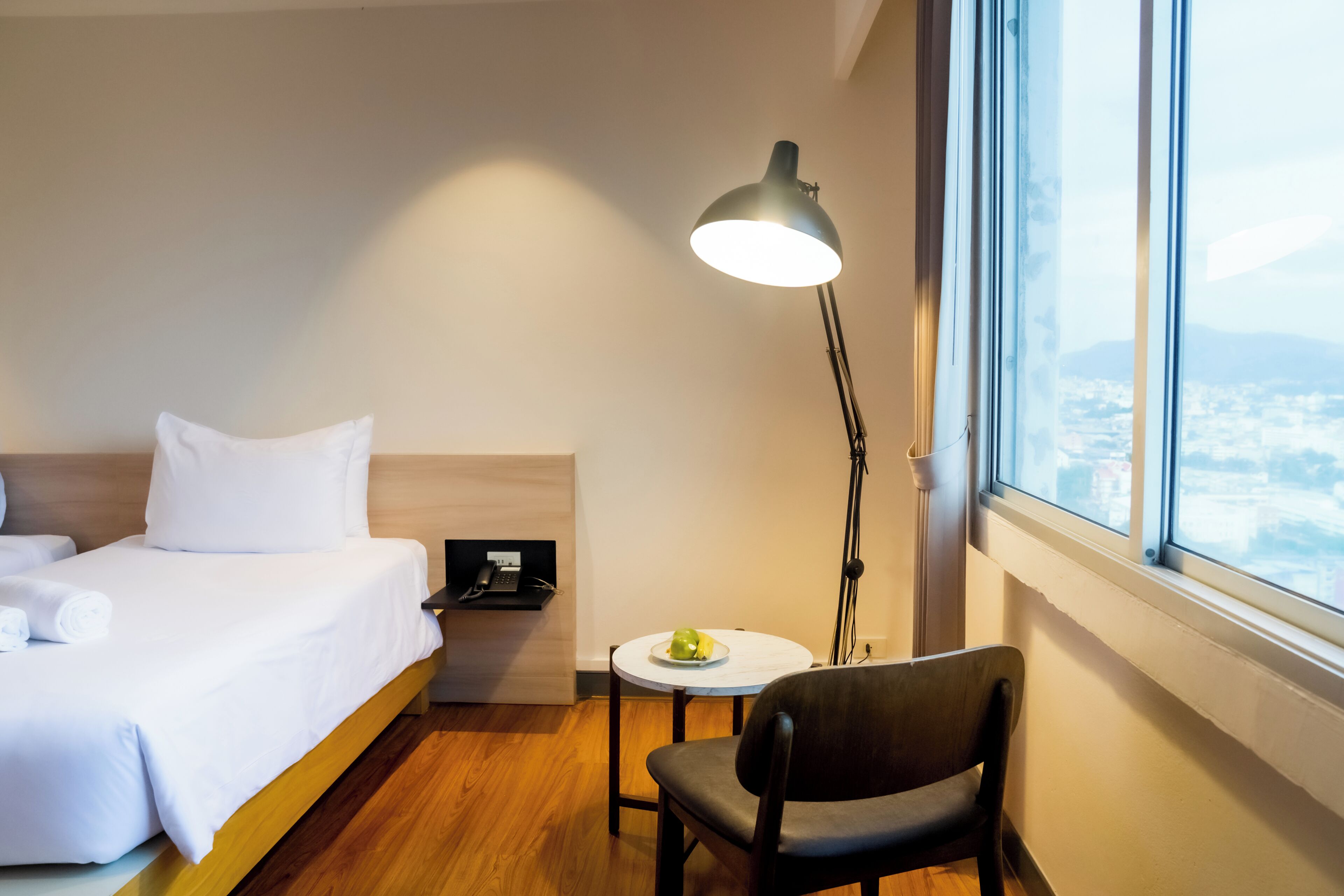 A hotel room with a bed, a desk with a lamp, a chair, and a window with a city view.