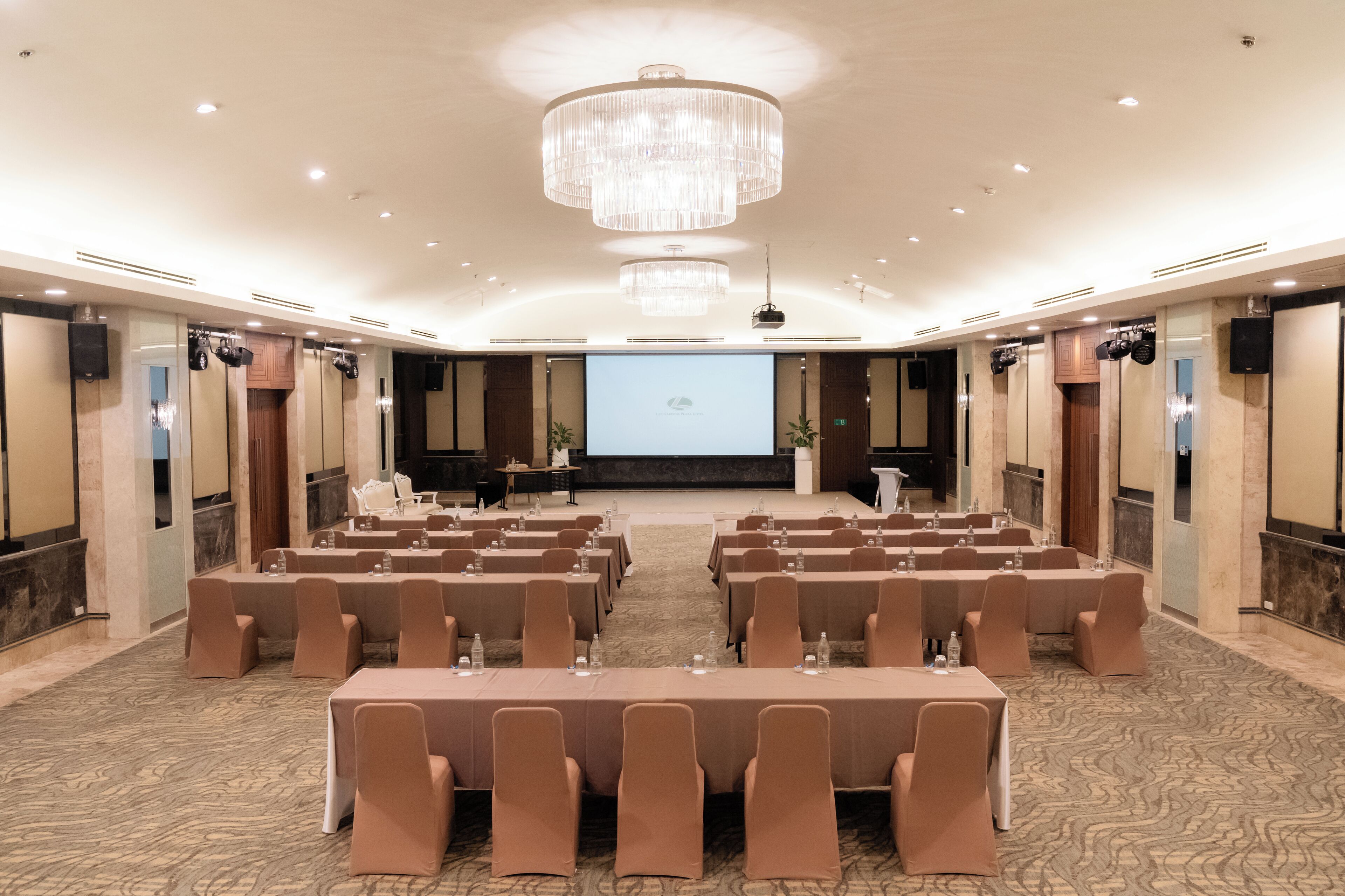 A conference room with rows of tables and chairs, a large screen at the front, and a chandelier.