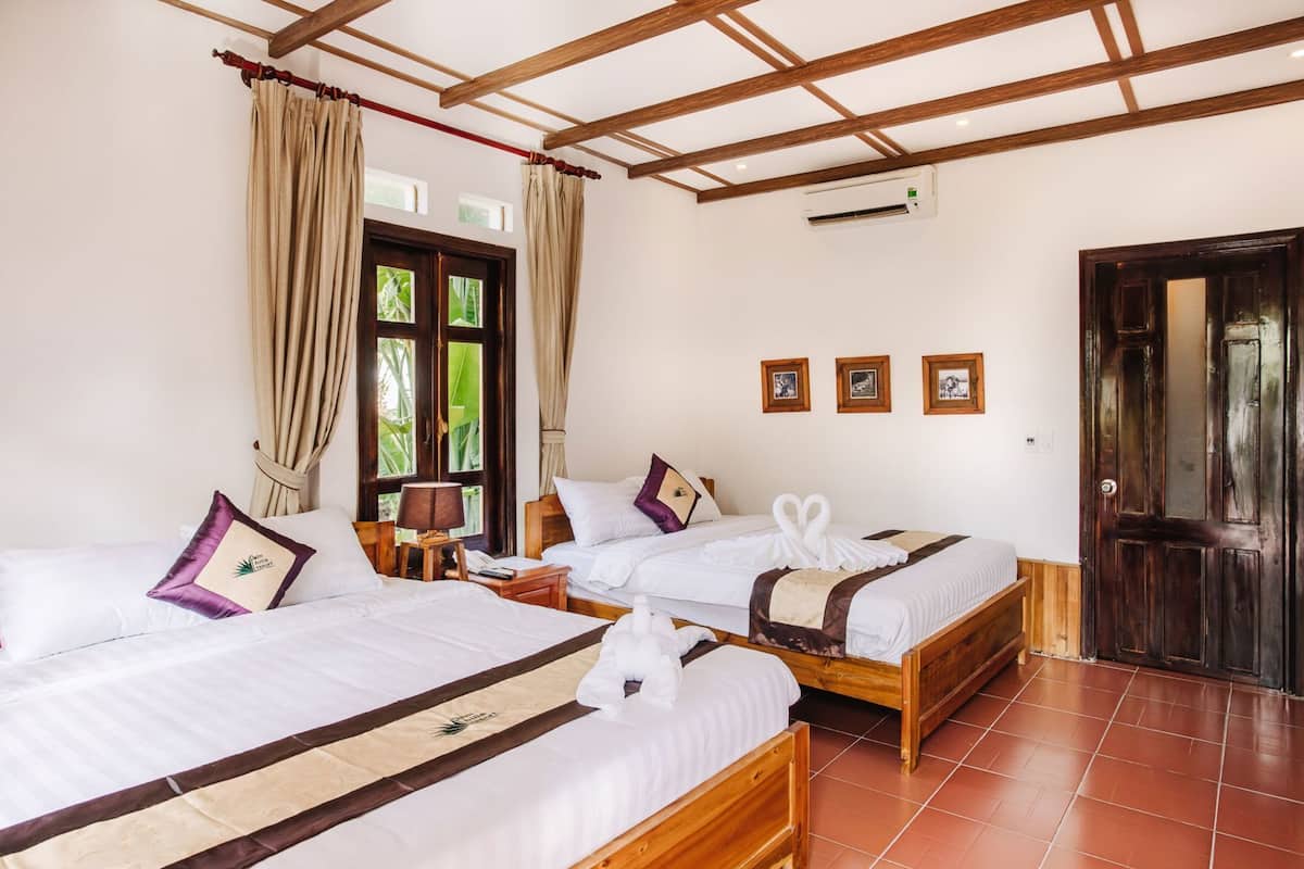 A hotel room with two wooden beds, a window with curtains, a door, and a wall-mounted air conditioner.
