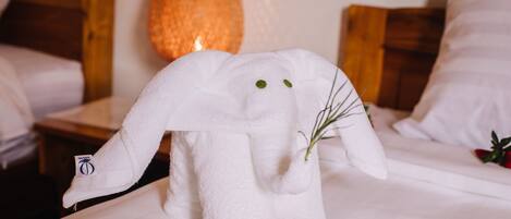 Una cama bien hecha con una toalla con forma de elefante, una flor roja y una lámpara al fondo.