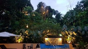 Una piscina con borde azul, rodeada de abundante vegetación y un muro de piedra con una fuente de agua. Hay sillones de descanso y una mesa cerca de la piscina.