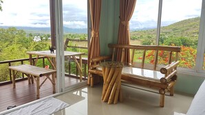 Una habitación con vista a un paisaje exuberante, un banco de madera, una mesa de vidrio y una silla de bambú.