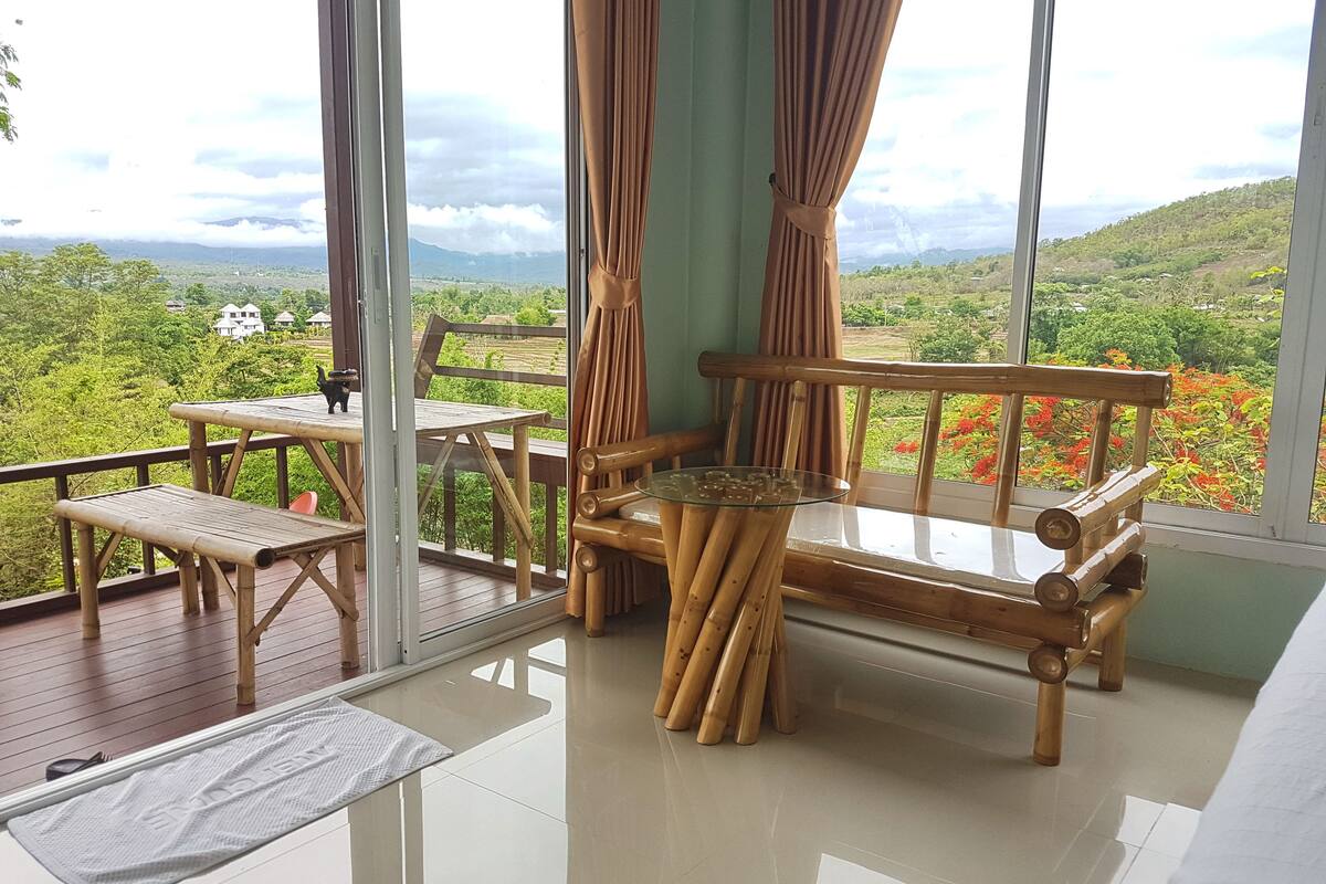 A room with a view of a lush landscape, a wooden bench, a glass table, and a bamboo chair.
