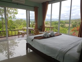 Un dormitorio con una cama grande, un balcón con mesa y sillas, y vistas a zonas verdes y montañas.