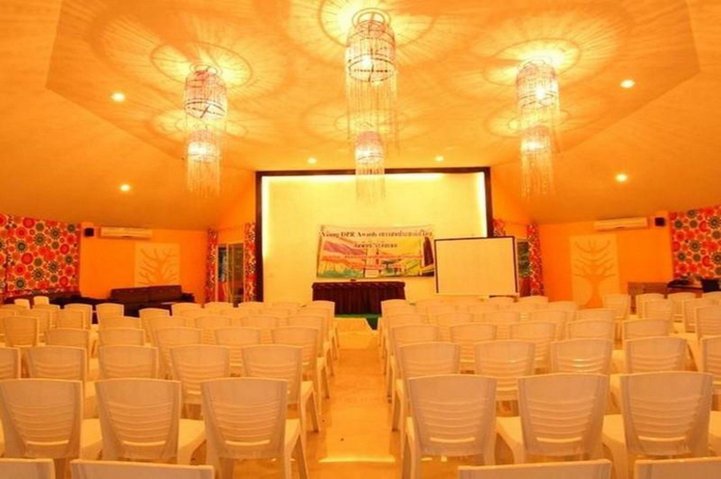 Uma sala de conferências com fileiras de cadeiras brancas, um palco com um banner e lustres decorativos.