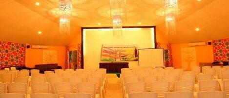 Uma sala de conferências com fileiras de cadeiras brancas, um palco com um banner e lustres decorativos.