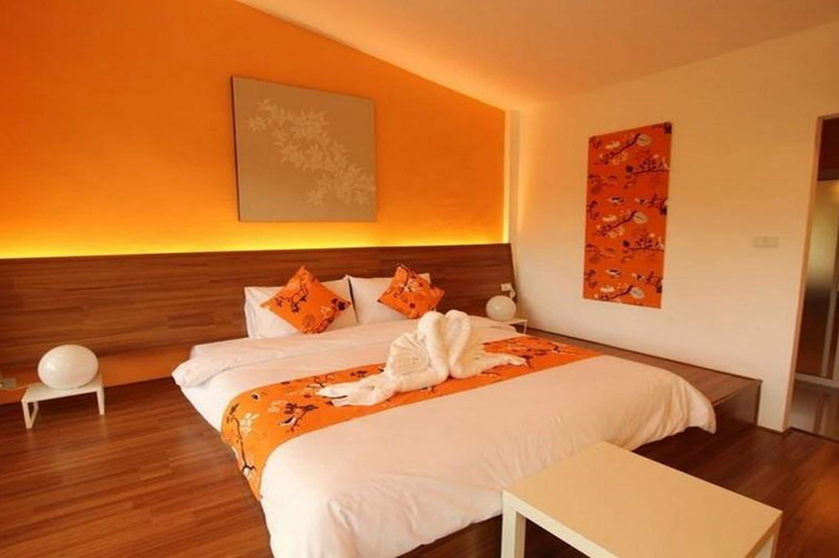 A modern hotel room with a wooden headboard, a large bed with orange bedding, and a white bedside table.
