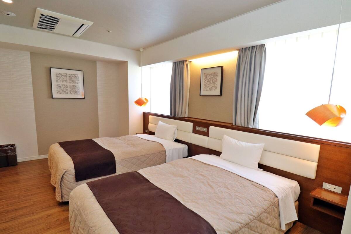 A hotel room with two beds, a wooden floor, a large window with curtains, and a ceiling air vent.