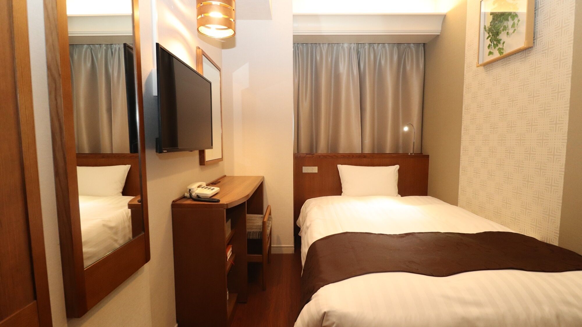 A hotel room with a bed, a desk with a telephone, a television mounted on the wall, and a window with curtains.