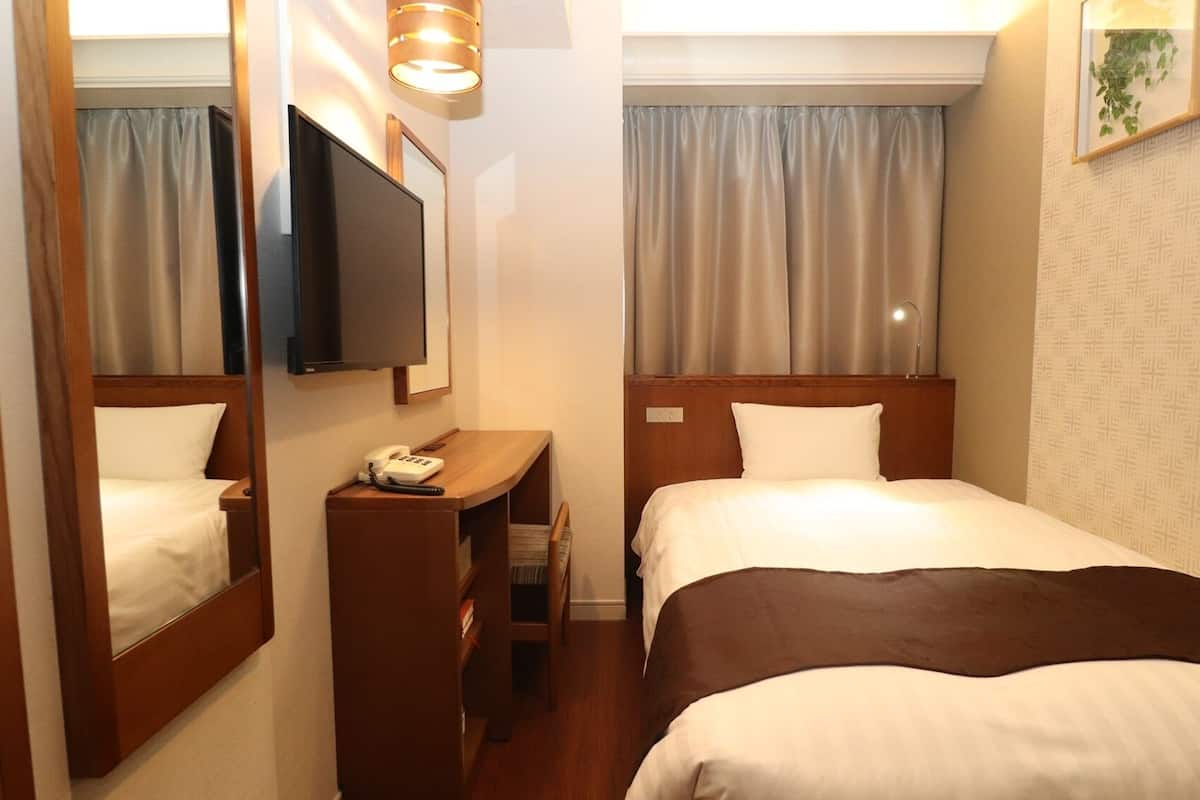 A hotel room with a bed, a desk with a telephone, a television mounted on the wall, and a window with curtains.