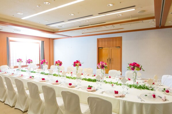 A conference room with a long table set for a formal event, equipped with chairs, tables, and floral centerpieces.