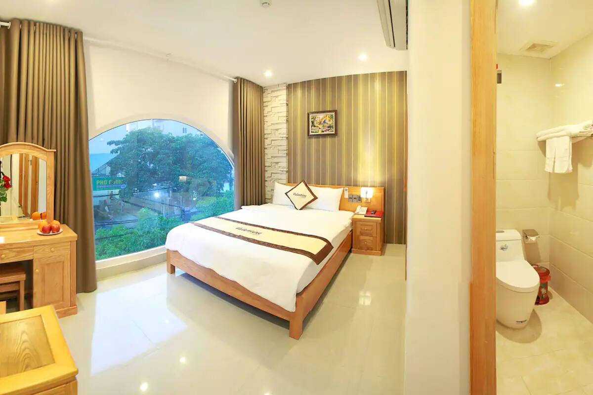 A hotel room with a large bed, a wooden dresser, a vanity with a mirror, and a view of greenery outside.
