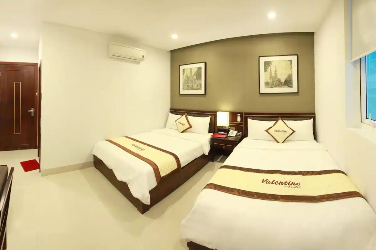A hotel room with two beds, each with a 'Valentine' themed bedspread, a bedside table with a lamp, and framed pictures on the wall.