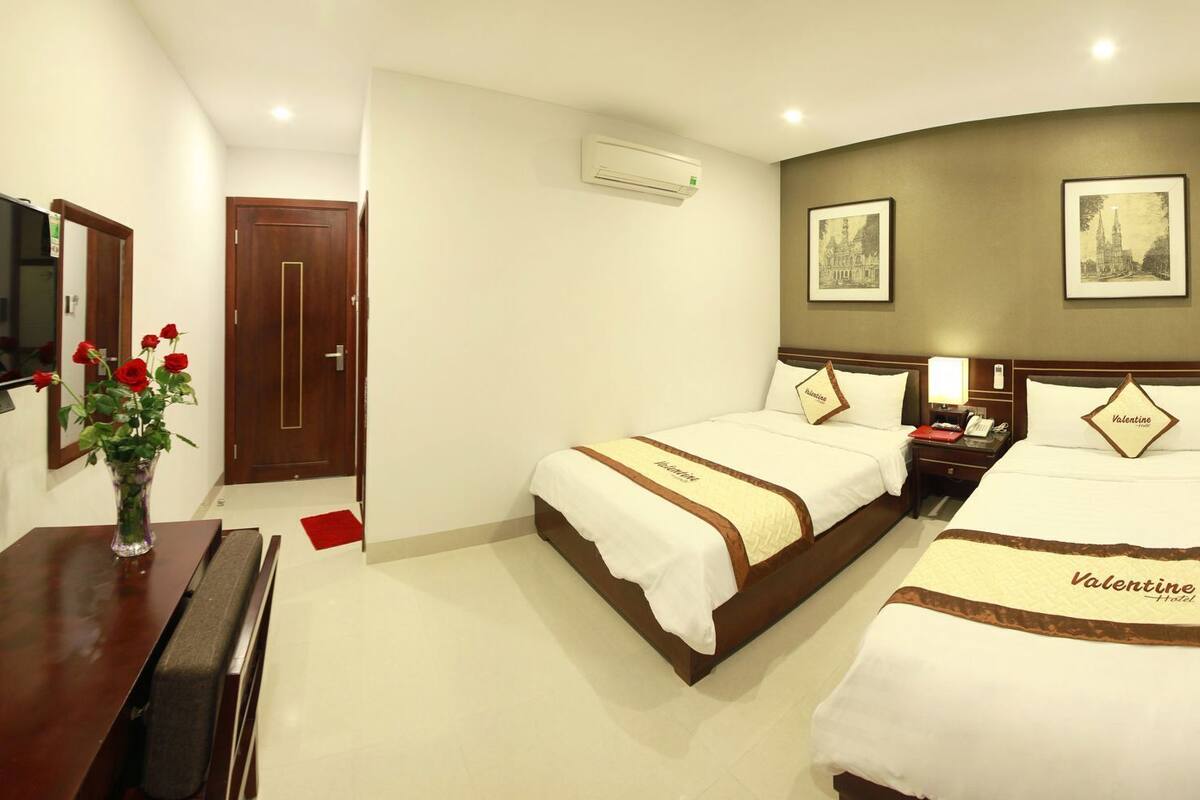 A hotel room with two beds, a desk, a TV, and framed pictures on the wall.
