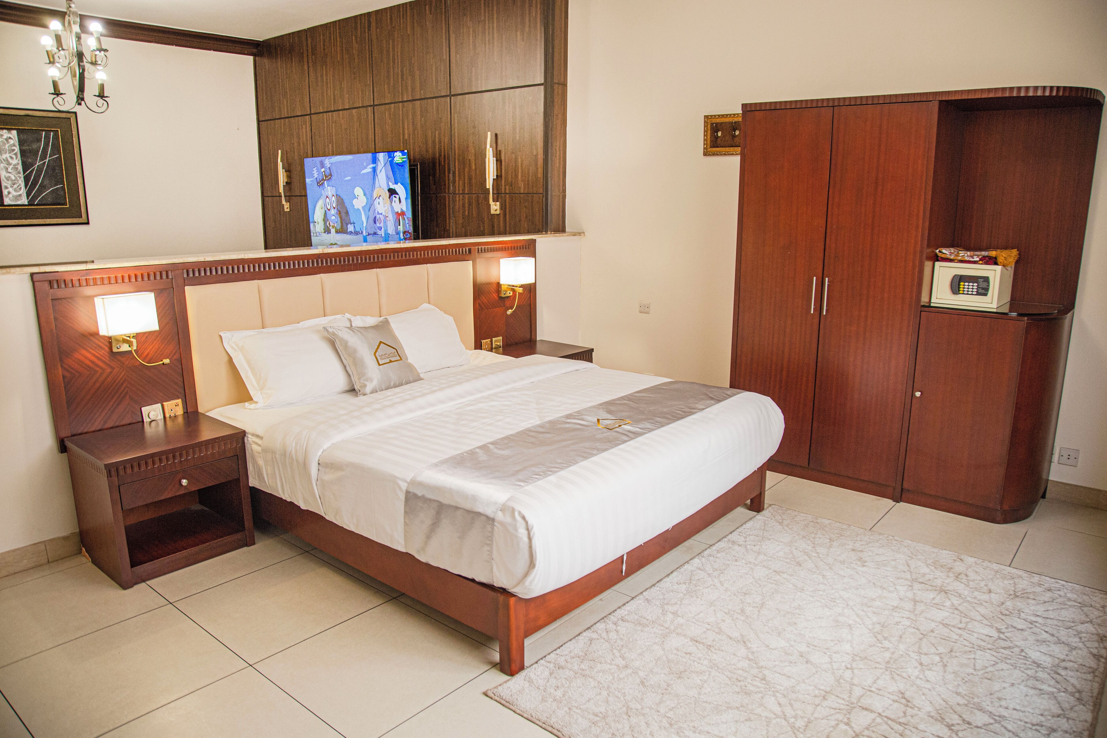 A hotel room with a large bed, wooden headboard, bedside tables with lamps, a wardrobe, and a television on the wall.