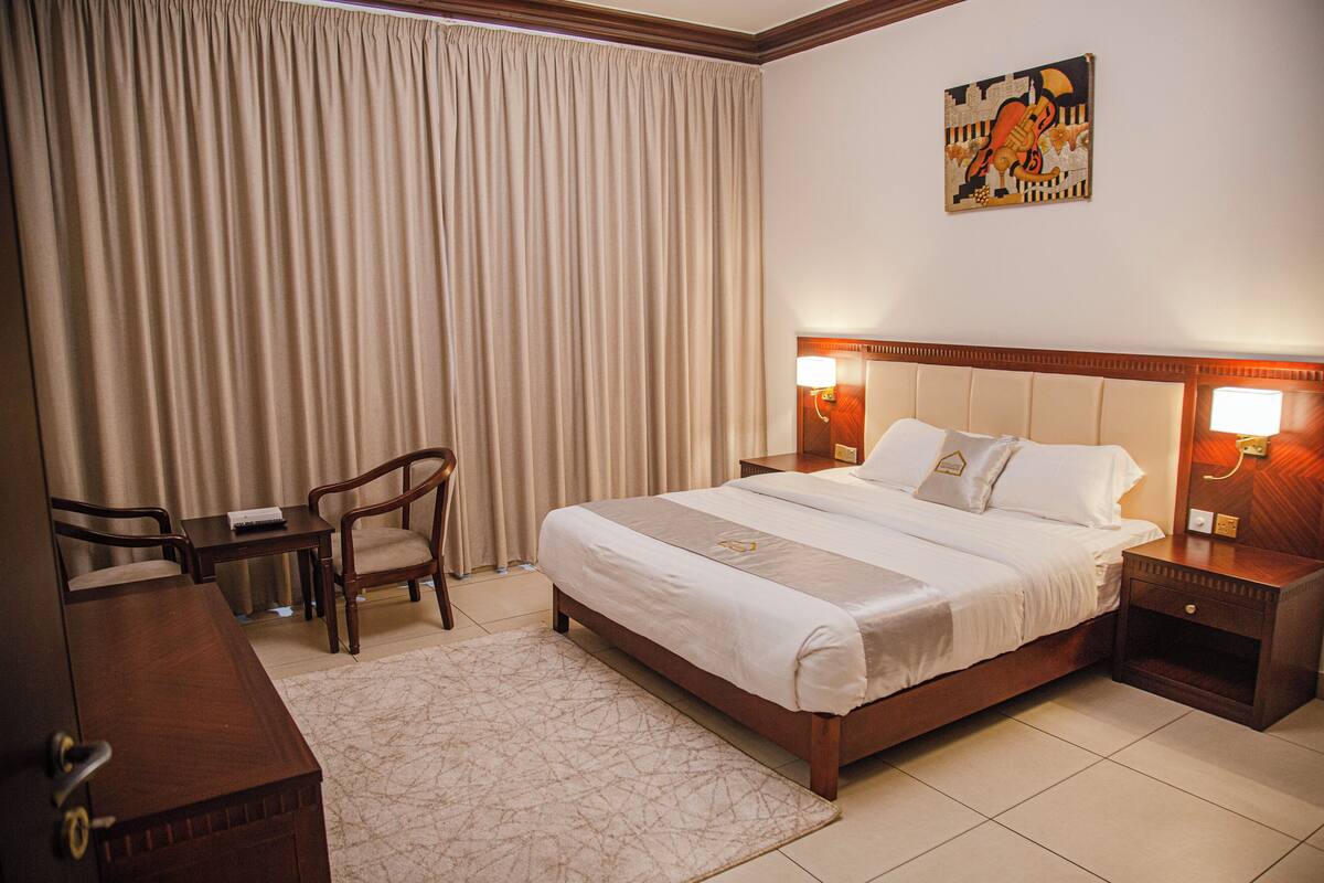 A hotel room with a large bed, a desk, a chair, and a nightstand with a lamp.