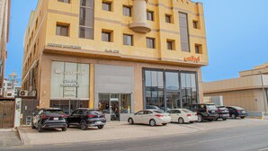 Un edificio de varios pisos con fachada amarilla, un amplio acceso de vidrio y varios automóviles estacionados adelante.