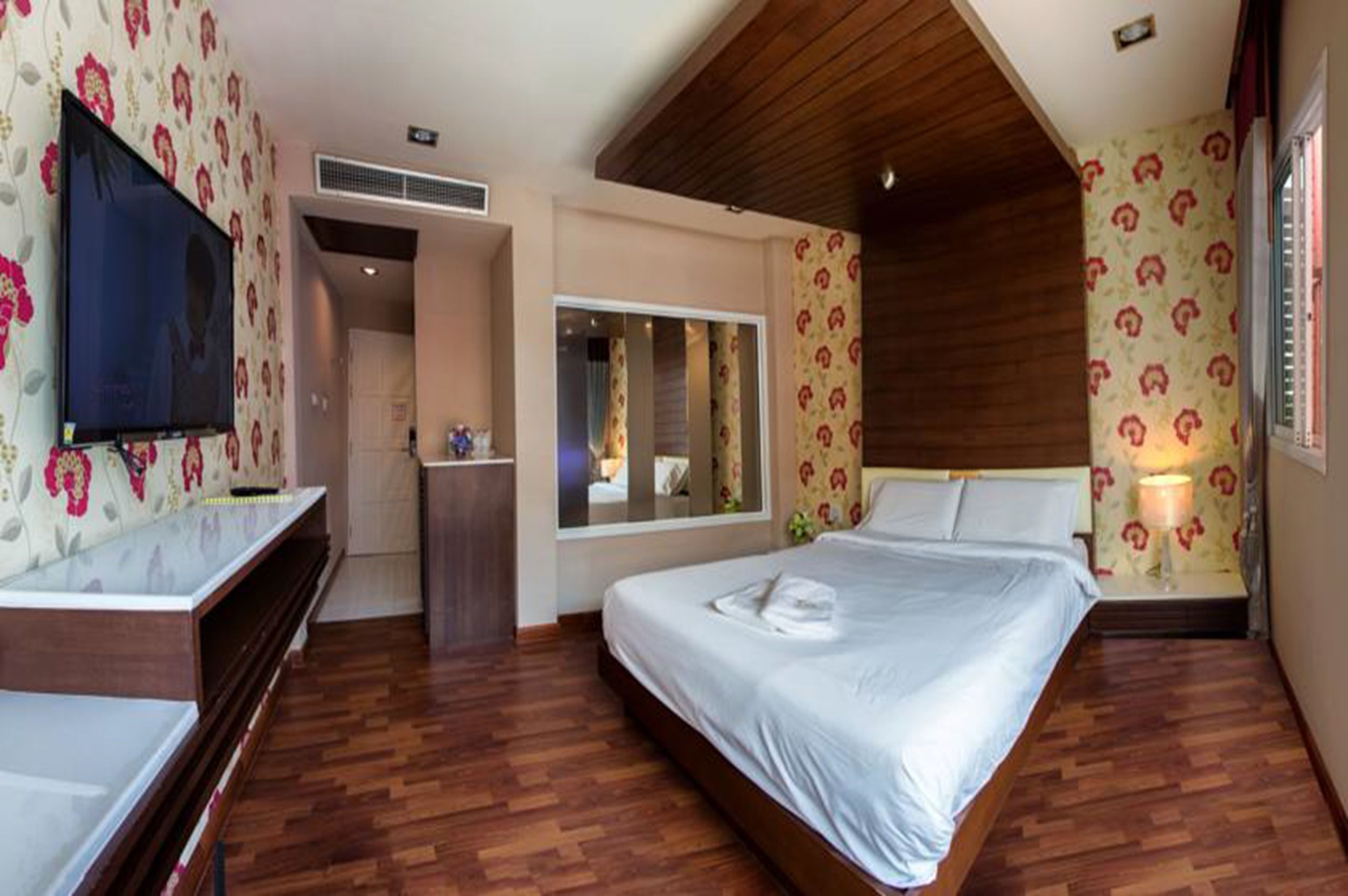 A bedroom with a wooden ceiling, a bed with white linens, a TV on a wooden stand, and floral wallpaper.