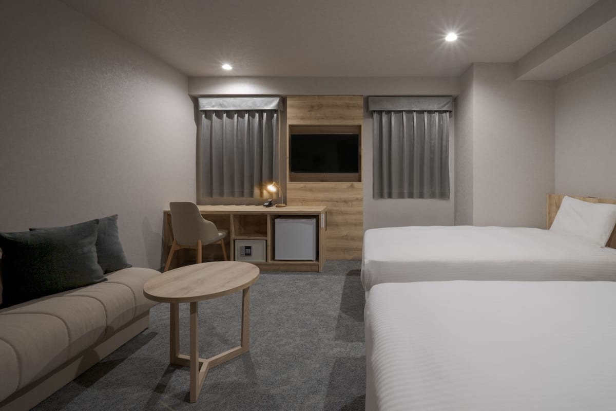A hotel room with a sofa, a small round table, a desk with a chair, and two beds.