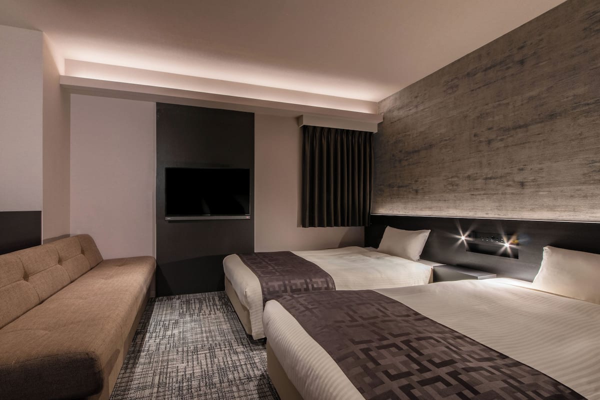 A modern hotel room with a sofa, two beds, a flat-screen TV, and a large wall-mounted light fixture.