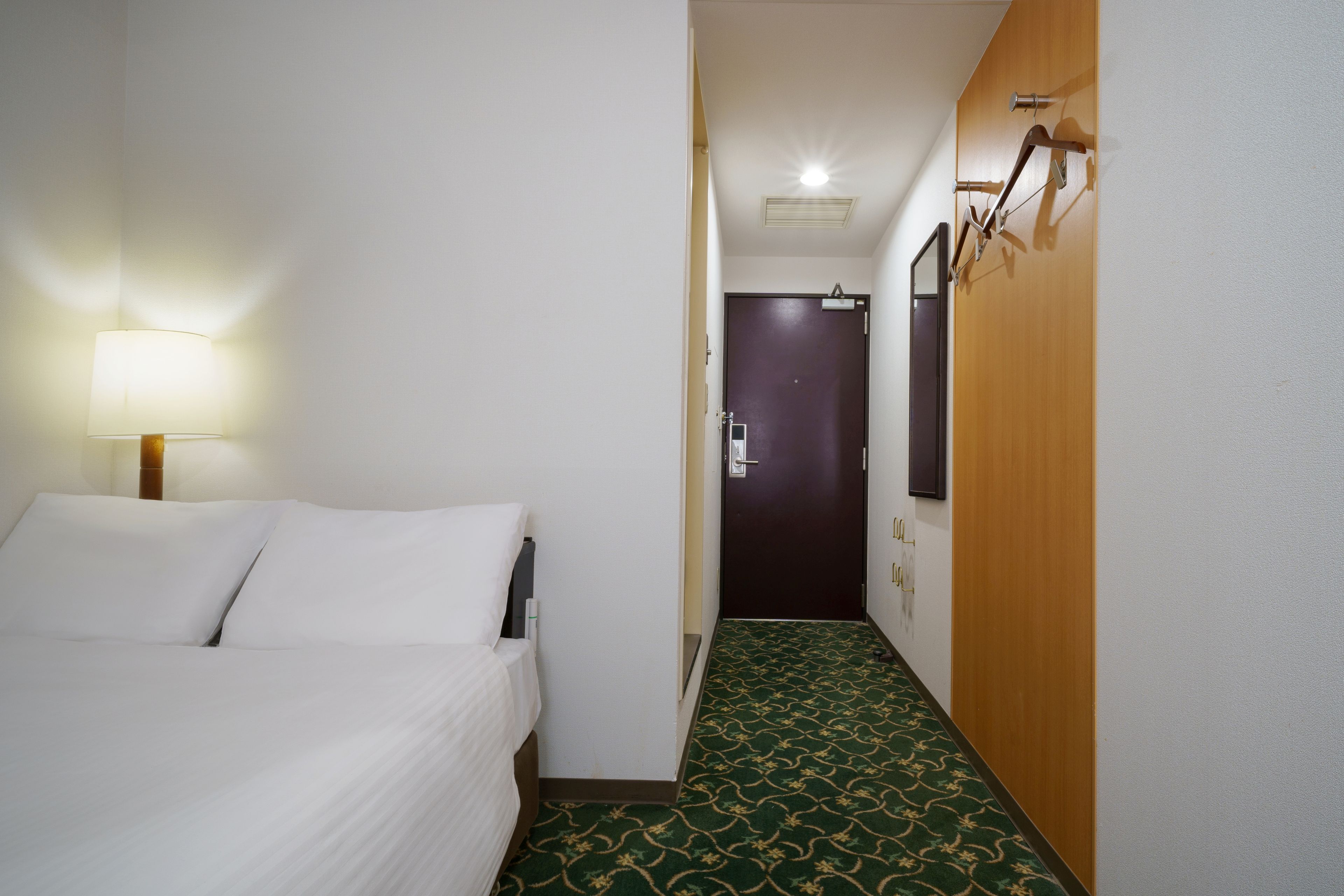 A hotel room with a bed, a lamp, a door, and a hallway with a patterned carpet.