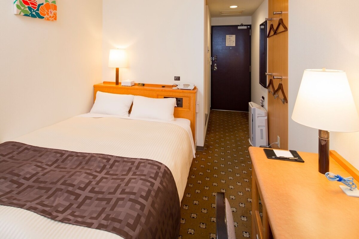 A hotel room with a bed, a desk with a lamp, a telephone, and a wall-mounted mirror.