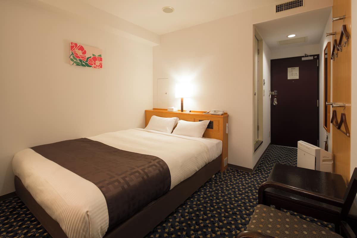 A hotel room with a bed, a desk, a chair, a lamp, and a door.