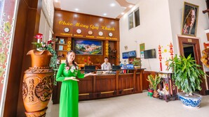 A hotel reception area with a wooden reception desk, a large vase with flowers, and a TV screen displaying a cityscape. - Hoang Duc Hotel (Chau Doc)