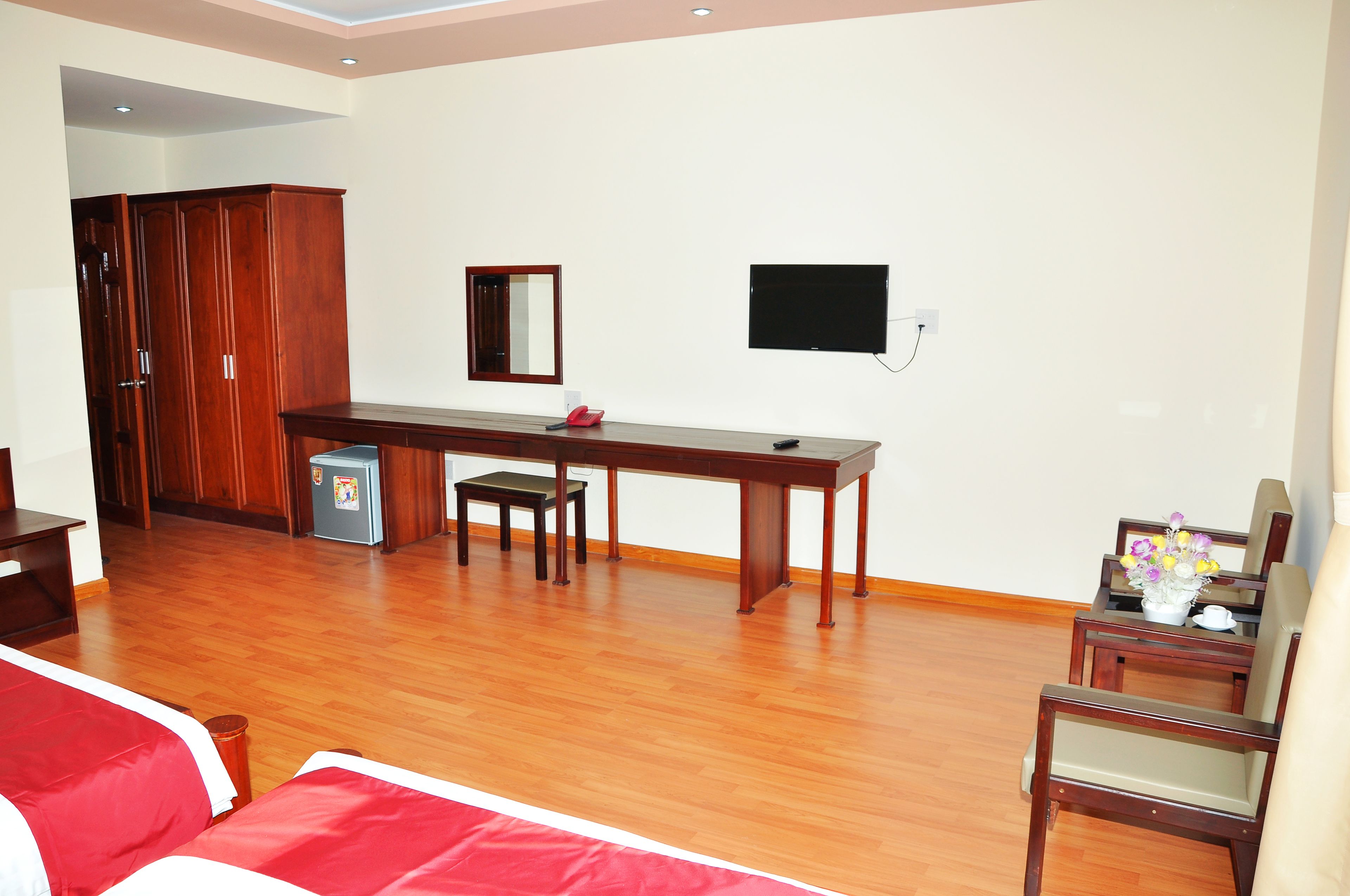 A hotel room with a wooden floor, a bed with red bedding, a wooden desk with a chair, a small refrigerator, a TV mounted on the wall, and a wardrobe.