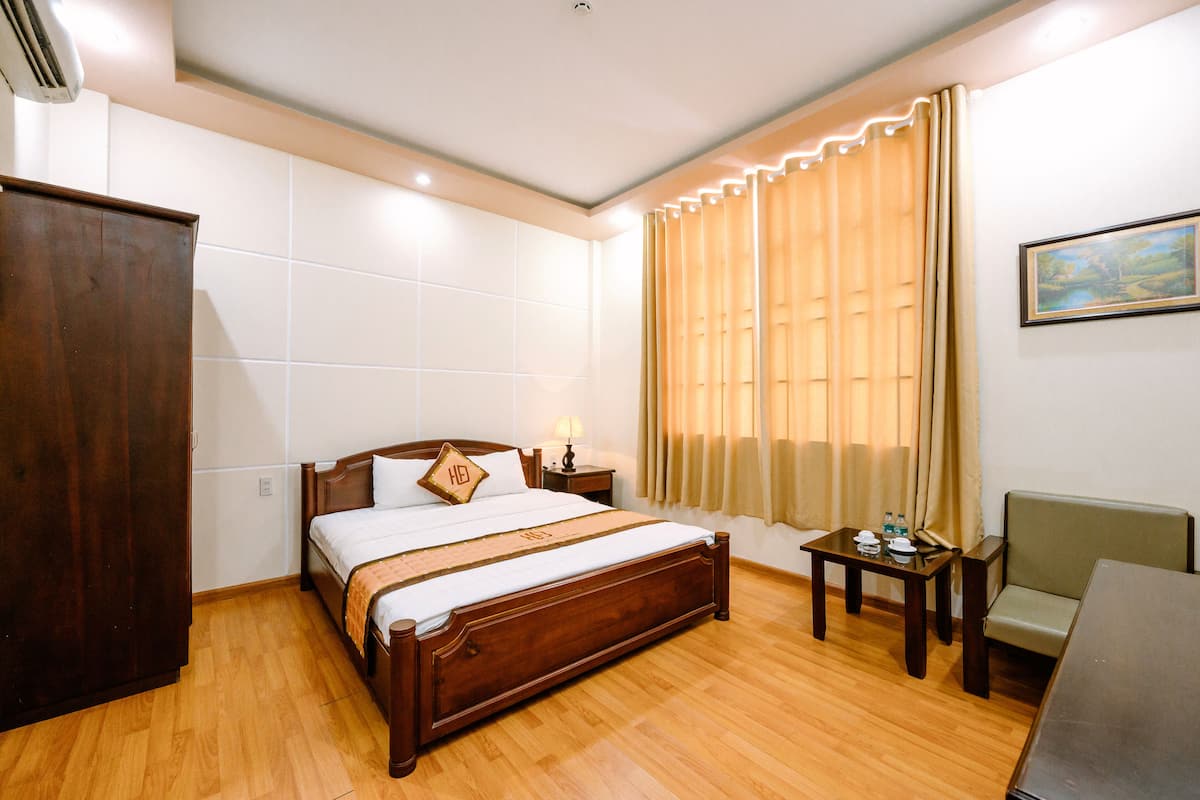 Habitación de hotel con una cama de madera, una mesita de noche con una lámpara, una pequeña mesa con una silla y un cuadro en la pared.