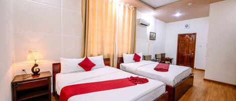 A hotel room with two beds, a wooden floor, a nightstand with a lamp, and a door.