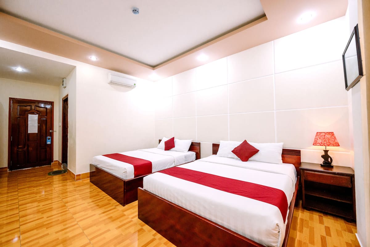 Habitación de hotel con dos camas, piso de madera, paredes blancas y una lámpara roja sobre una mesita de noche de madera.