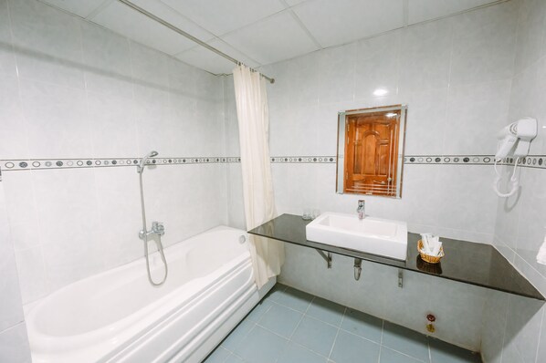 A bathroom with a bathtub, a glass vanity with a sink, a hairdryer, and a showerhead. - Hoang Duc Hotel (Chau Doc)
