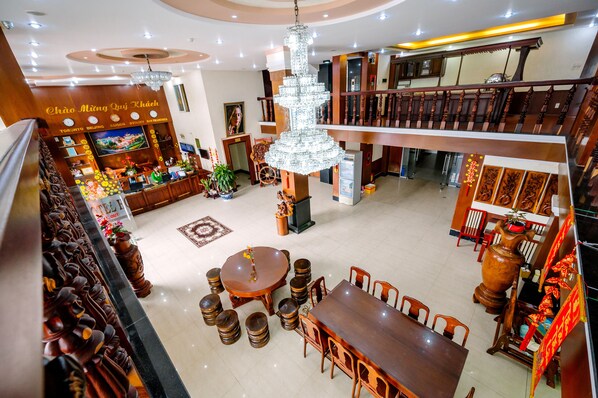 A spacious lobby with a high ceiling, a chandelier, a traditional wooden reception desk, and a dining area with a round table and chairs.