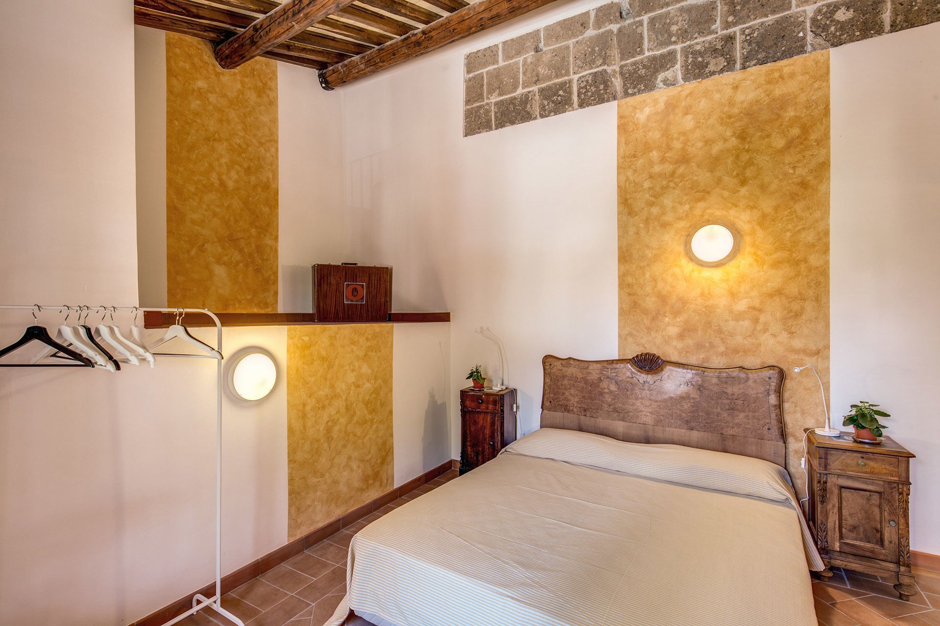 Un dormitorio con cama, cabecera de madera, estante en la pared, perchero y mesita de noche con lámpara y planta.